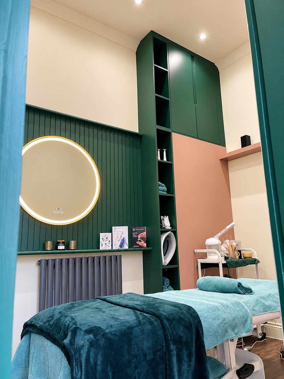 Treatment room with a full-height green storage unit, fluted panelling and a round lit mirror
