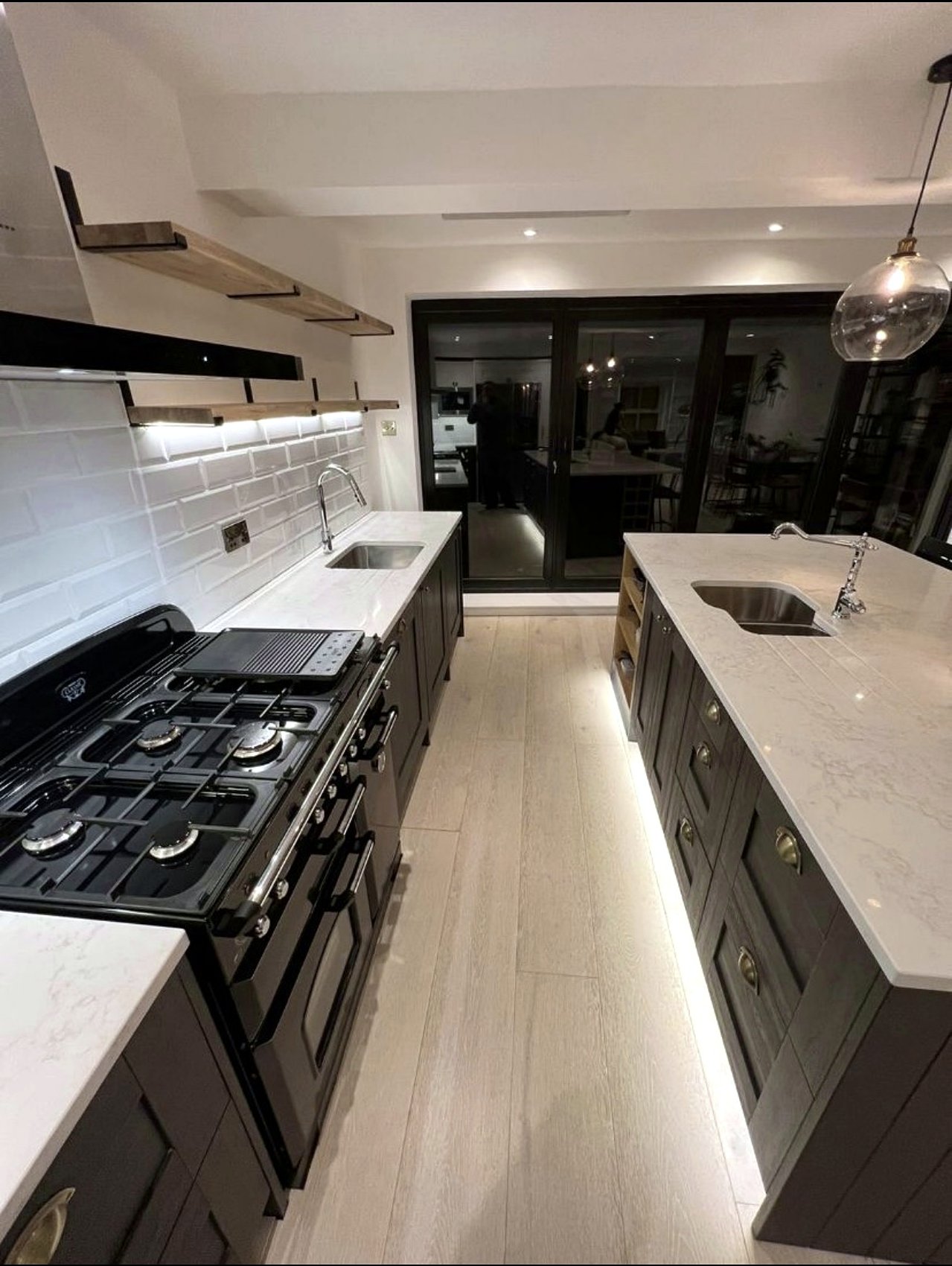 Dark shaker kitchen with a black range cooker, pale stone worktops, oak shelves and lit plinths